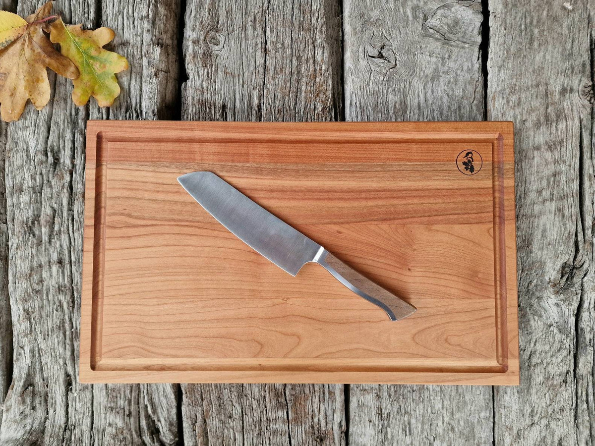 Schneidebrett mit Saftrille aus Kirschbaum auf alten Eichenbalken. Dekoriert mit einem Kochmesser von Güde und Eichenblättern.
