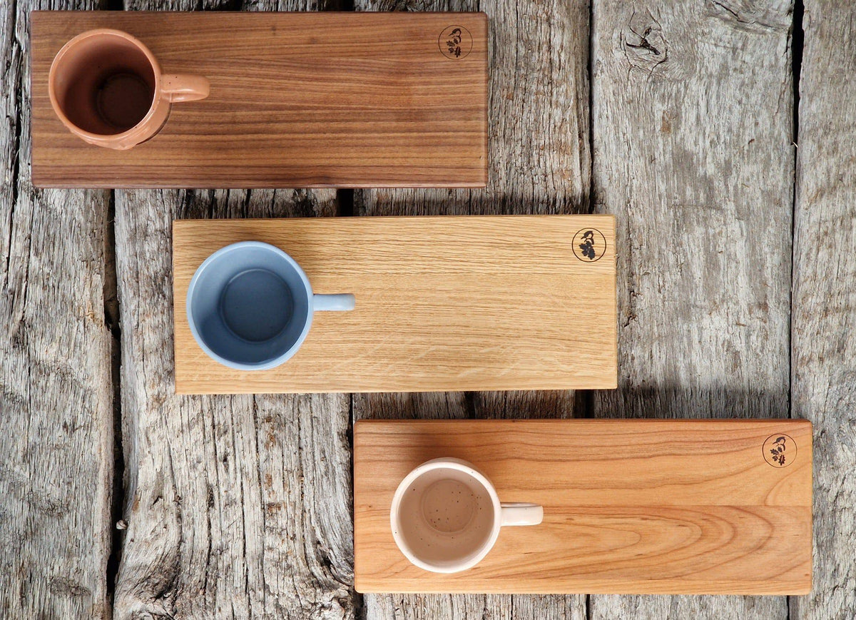 Rechteckige Holz Tabletts in Eiche, Kirsche und Nussbaum, jeweils mit einer Vertiefung für eine große Tasse. Dekoriert mit 3 Bechern mit Gesicht. Auf einem Eichenbalken.