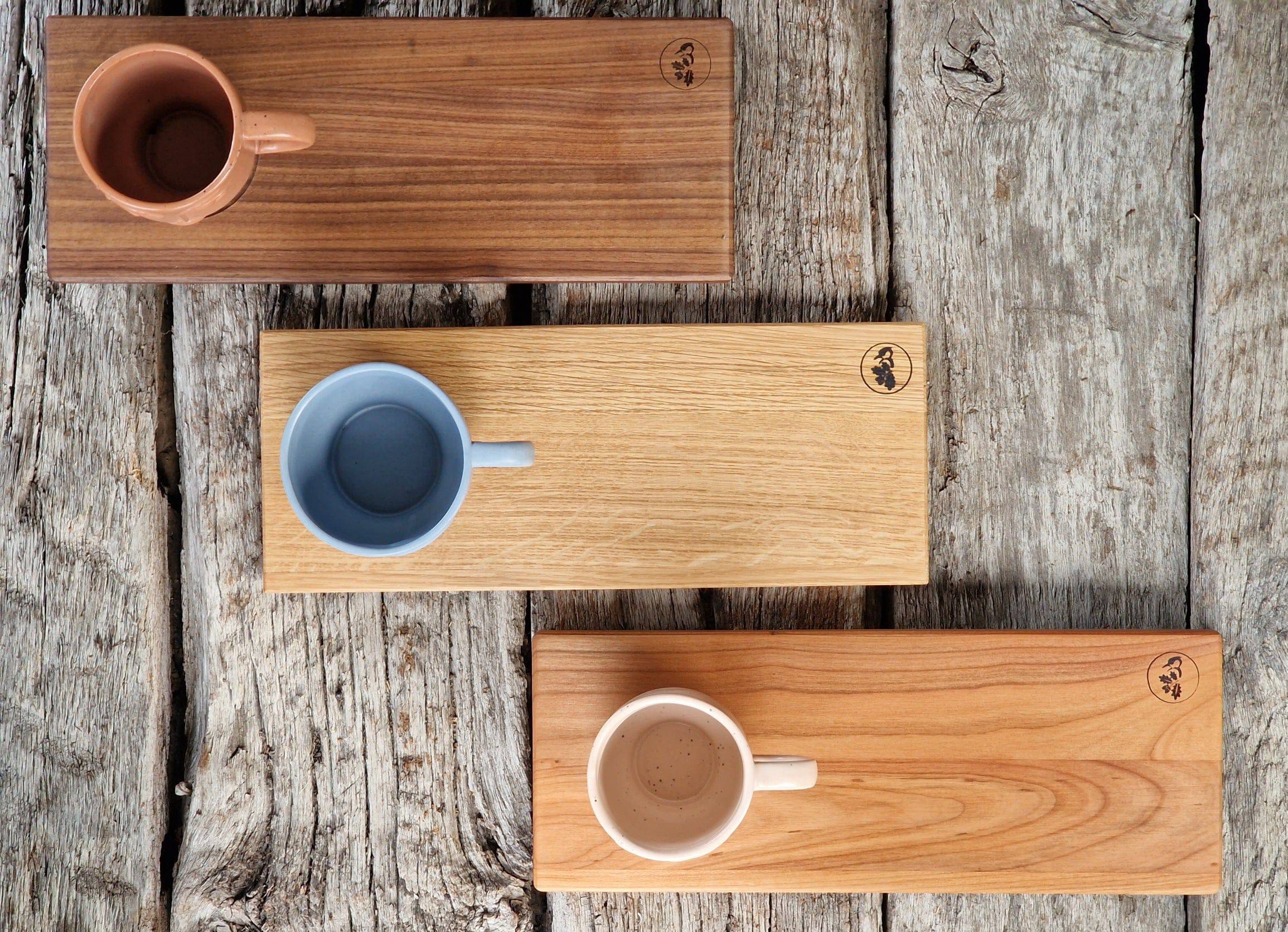 Rechteckige Holz Tabletts in Eiche, Kirsche und Nussbaum, jeweils mit einer Vertiefung für eine große Tasse. Dekoriert mit 3 Bechern mit Gesicht. Auf einem Eichenbalken. 